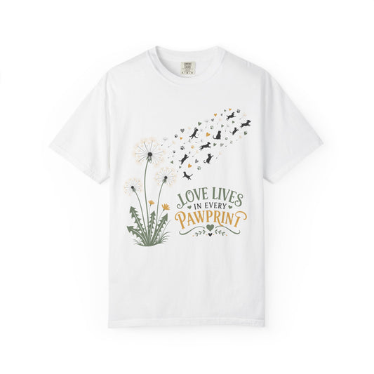 A white short-sleeve crewneck t-shirt featuring a cute graphic of dandelions, a puff of black and orange birds, and the text 'LOVE LIVES IN EVERY PAWPRINT' on the front.