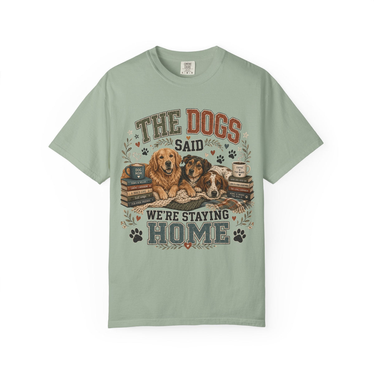 A sage green short-sleeve t-shirt with a graphic print of two dogs sitting on a bed with books, accompanied by the text 'THE DOGS SAID WE'RE STAYING HOME' and paw prints.