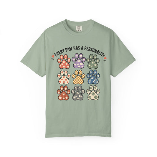 A sage green short-sleeve t-shirt with a graphic print of nine colorful dog paw prints and the text 'EVERY PAW HAS A PERSONALITY' on a white background.
