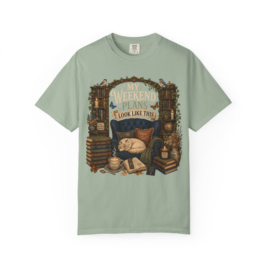 A sage green short-sleeve t-shirt with a detailed graphic print on the front. The graphic features a cozy scene with a sleeping cat in a blue armchair, surrounded by books, a coffee cup, and a sign that reads 'MY WEEKEND PLANS LOOK LIKE THIS'.