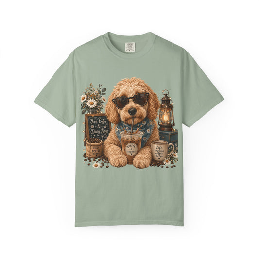 A sage green short-sleeve t-shirt featuring a central graphic illustration of a fluffy golden-brown dog wearing sunglasses, surrounded by coffee cups, a lantern, and flowers.
