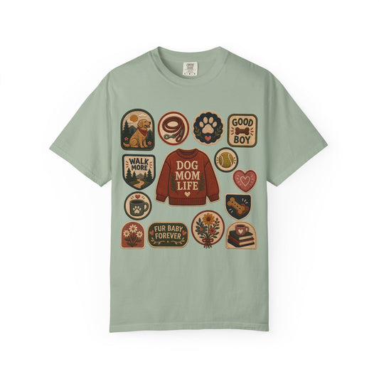 A sage green short-sleeve t-shirt with a graphic print of various dog-themed patches surrounding a central red 'Dog Mom Life' sweater, displayed on a clean white background.
