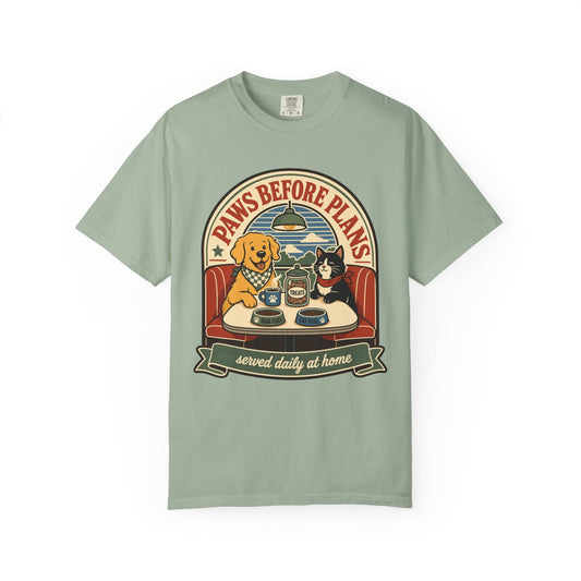 A sage green short-sleeve t-shirt with a vintage-style graphic on the chest featuring a golden retriever and a black cat sitting at a diner table under the text 'PAWS BEFORE PLANS' and 'served daily at home'.