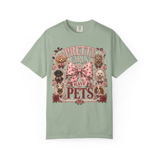 A sage green short-sleeve t-shirt with a vintage-style graphic print featuring the text 'PRETTY GIRLS HAVE PETS' in pink and red, surrounded by illustrations of various dog breeds and a pink polka-dot bow.