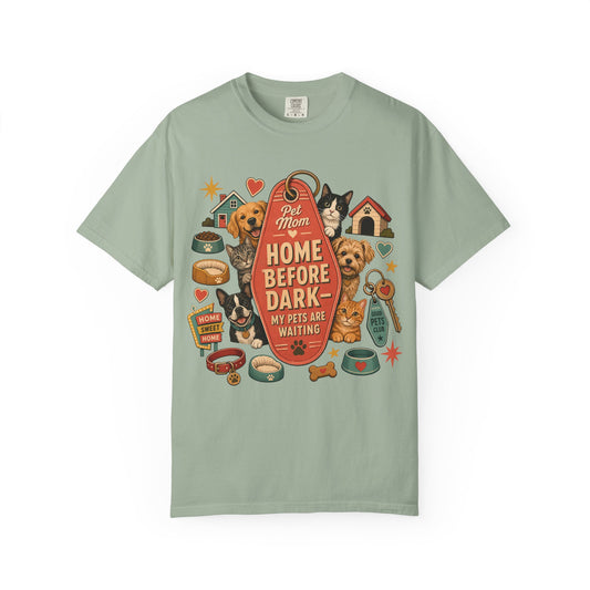 A sage green short-sleeve t-shirt with a colorful graphic print featuring various dog breeds, pet accessories, and a central red tag with the text 'Pet Mom HOME BEFORE DARK - MY PETS ARE WAITING'.