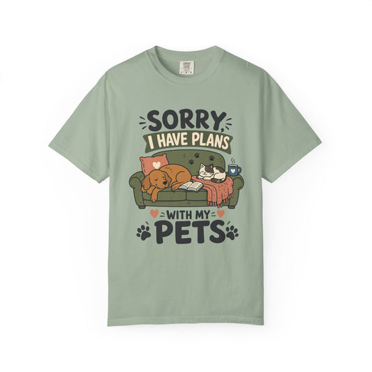 A sage green short-sleeve t-shirt with a cartoon graphic of a dog and cat on a couch, accompanied by the text 'SORRY I HAVE PLANS WITH MY PETS'.