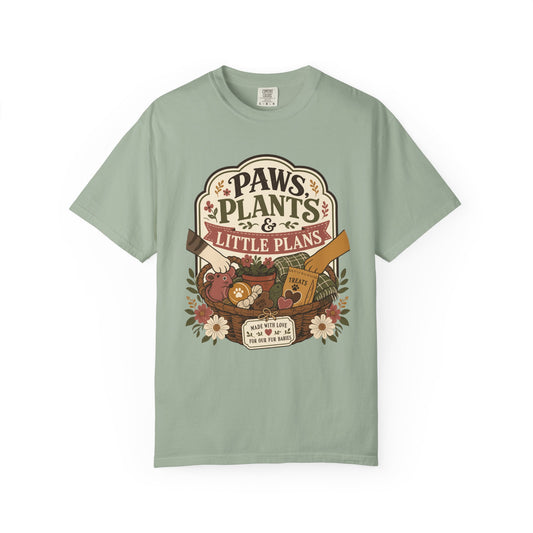 A sage green short-sleeve t-shirt with a vintage-style graphic on the chest that reads 'PAWS, PLANTS & LITTLE PLANS' above a whimsical illustration of a basket, flowers, and a small dog.