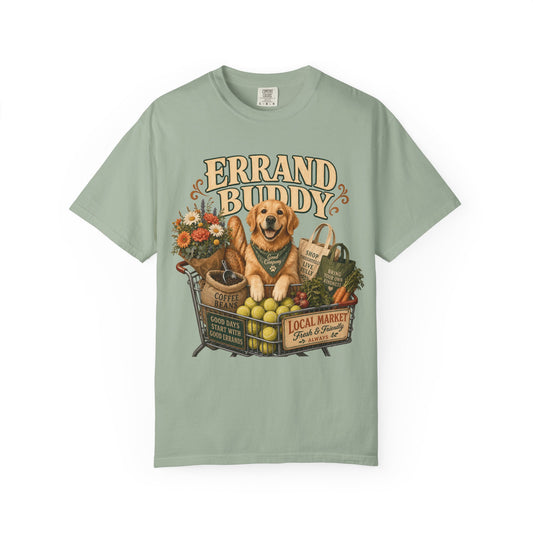 A sage green short-sleeve t-shirt with a vintage-style graphic of a golden retriever dog sitting in a market cart surrounded by fruits, vegetables, flowers, and shopping bags. The text 'ERRAND BUDDY' is printed above the illustration.