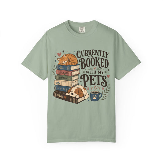 A sage green short-sleeve t-shirt with a graphic print of a cat on a stack of books, a sleeping puppy, a coffee cup, and the text 'CURRENTLY BOOKED WITH MY PETS'.