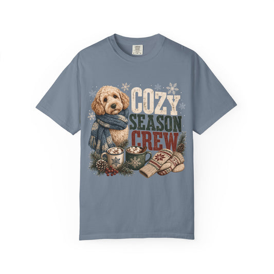 A slate blue crewneck t-shirt featuring a cozy holiday graphic with a golden doodoodle wearing a blue scarf, hot cocoa mugs, knit mittens, pinecones, and the text 'COZY SEASON CREW'.