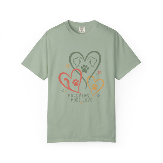 A sage green short-sleeve crewneck t-shirt featuring a graphic of three interlocking hearts in red, orange, and dark green, each containing a dog paw print, with the text 'MORE PAWS, MORE LOVE' printed below.