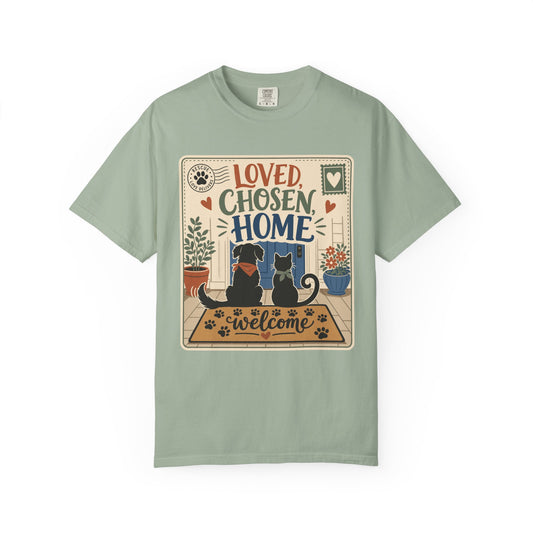 A sage green short-sleeve t-shirt with a central graphic print featuring the text 'LOVED CHOSEN HOME' above an illustration of a black dog and a black cat sitting on a 'Welcome' doormat.