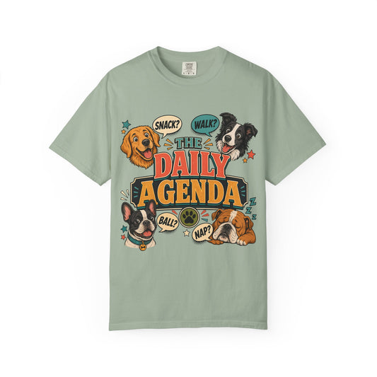A sage green short-sleeve t-shirt with a colorful graphic print featuring cartoon dogs and the text 'THE DAILY AGENDA'.