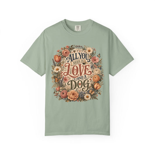 A sage green short-sleeve t-shirt with a vintage-style floral graphic on the chest that reads 'ALL YOU NEED IS LOVE AND A DOG'.