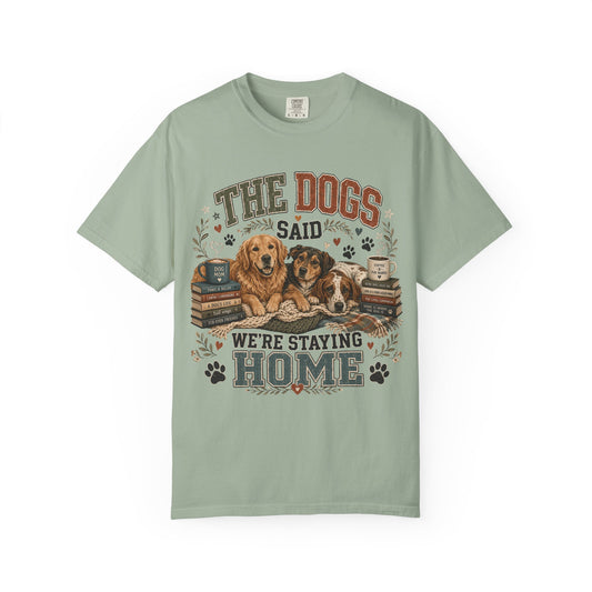 A sage green short-sleeve t-shirt with a graphic print of two dogs sitting on a bed with books, accompanied by the text 'THE DOGS SAID WE'RE STAYING HOME' and paw prints.