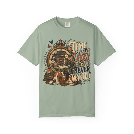 A sage green short-sleeve t-shirt with a vintage-style graphic on the front featuring a sleeping basset hound, a clock, and the text 'TIME SPENT COZY IS NEVER WASTED'.