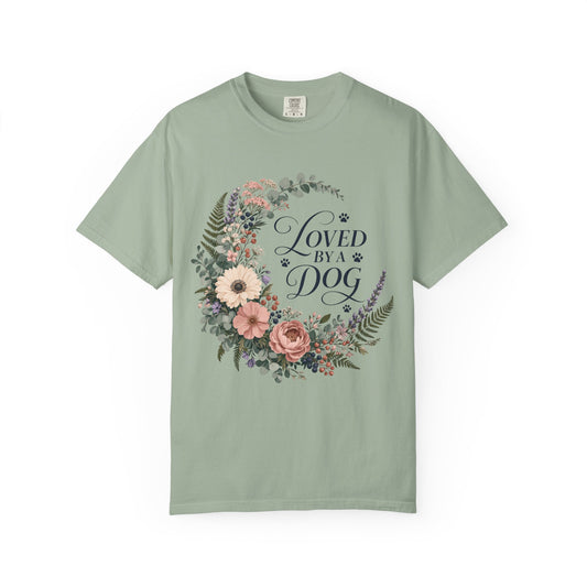 A sage green short-sleeve crewneck t-shirt featuring a central graphic of a floral wreath with the text 'LOVED BY A DOG' in a dark cursive font, displayed on a clean white background.