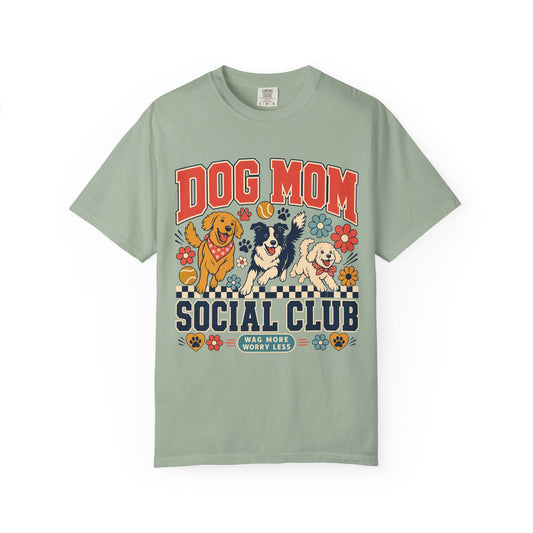 A sage green short-sleeve t-shirt with a colorful graphic on the front featuring three dogs, flowers, and the text 'DOG MOM SOCIAL CLUB' and 'WAS MORE WOOFY THAN'.
