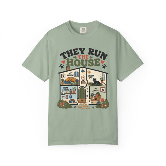 A sage green short-sleeve t-shirt with a detailed graphic on the front depicting a house with pets and the text 'THEY RUN THE HOUSE'.