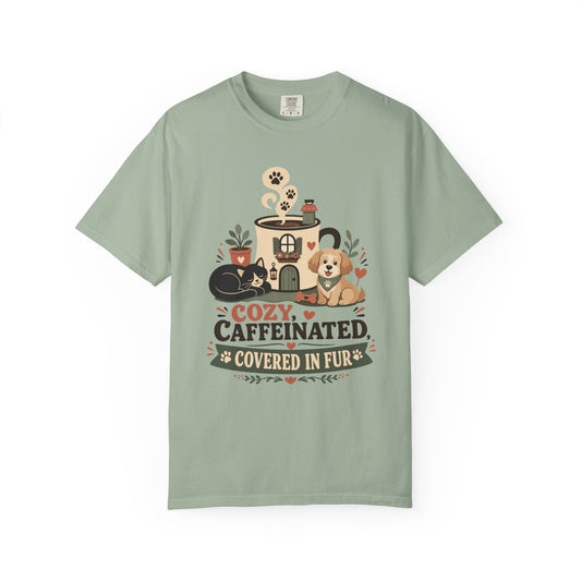 A sage green short-sleeve t-shirt with a cozy pet-themed graphic on the front. The graphic features a coffee mug with a paw print, a sleeping black cat, a golden retriever dog, and the text 'COZY, CAFFEINATED.