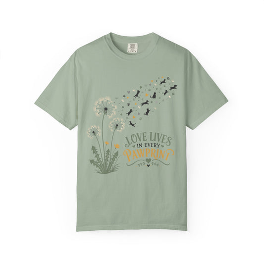 A sage green short-sleeve t-shirt with a graphic print of dandelions, flying dogs, and the text 'LOVE LIVES IN EVERY PAWPRINT'.