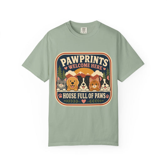 A sage green short-sleeve t-shirt with a retro-style graphic on the chest that reads 'PAWPRINTS WELCOME HERE HOUSE FULL OF PAWS' above a row of illustrated dogs, shown on a plain white background.
