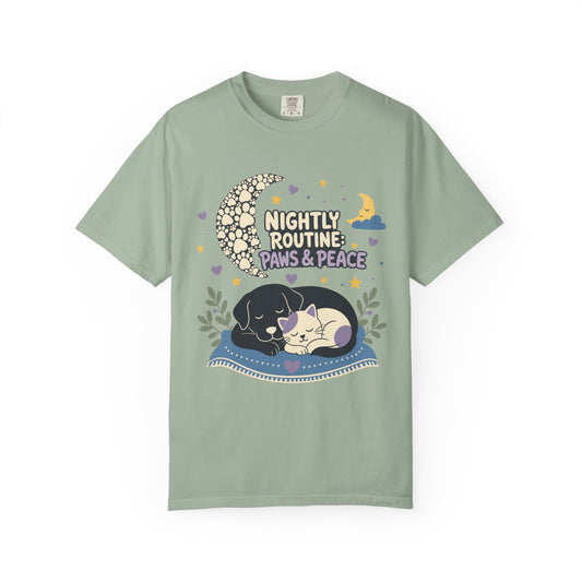 A sage green short-sleeve t-shirt with a cute graphic of a black and white puppy sleeping on a blue blanket under a crescent moon and stars, with the text 'Nightly Routine: Paws & Peace'.