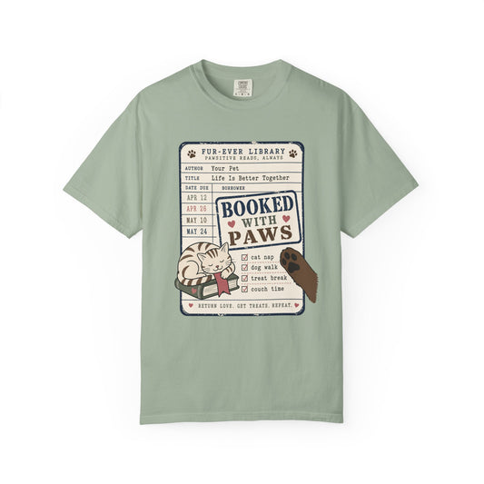 A sage green crewneck t-shirt featuring a 'Booked with Paws' graphic print on the front, displayed against a solid white background.