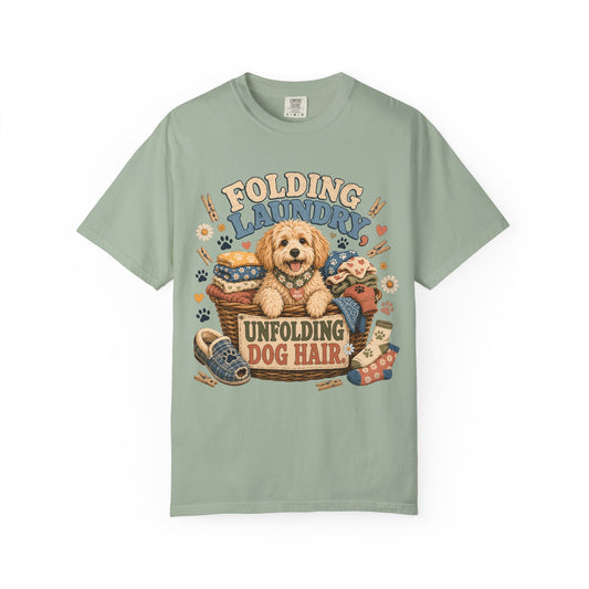 A sage green short-sleeve t-shirt with a colorful graphic of a fluffy dog and the text 'FOLDING LAUNDRY UNFOLDING DOG HAIR' on the front, displayed against a plain white background.