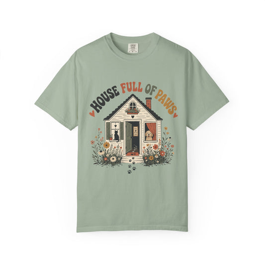 A sage green short-sleeve t-shirt with a graphic of a cozy house surrounded by flowers and the text 'HOUSE FULL OF PAWS' in orange and dark green lettering.